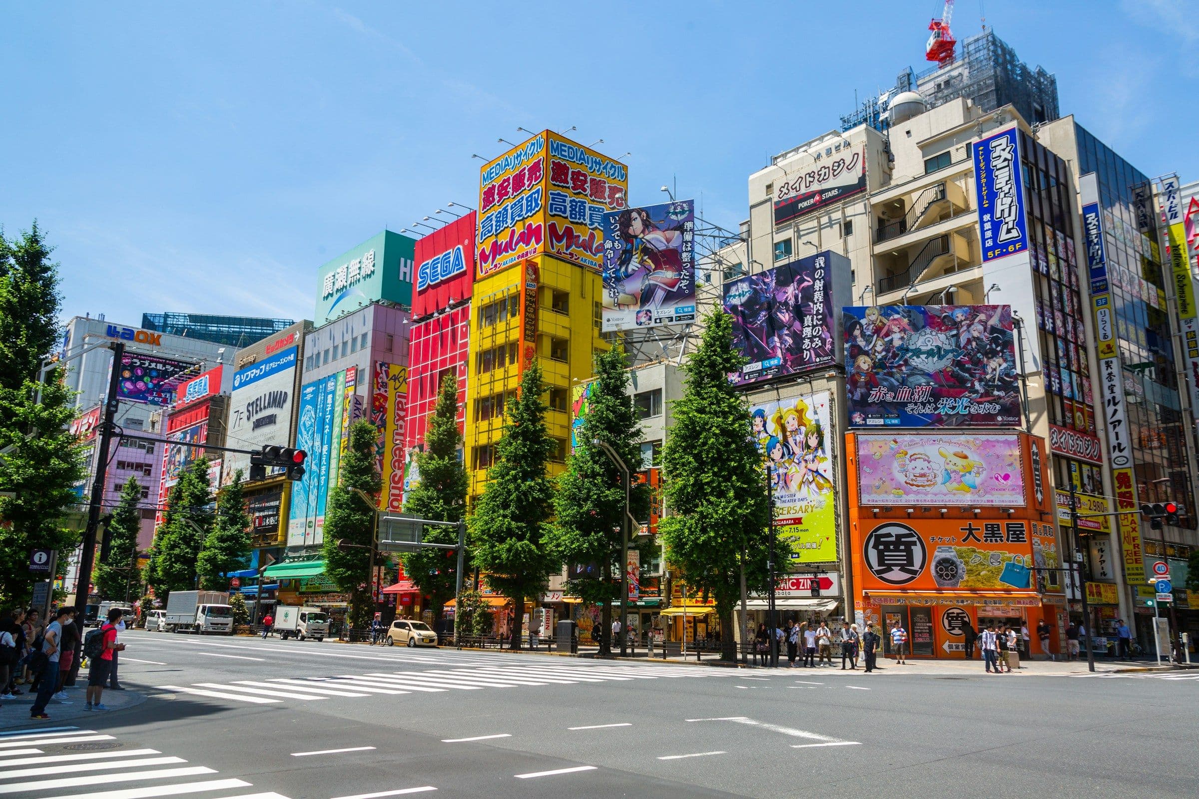 A busy intersection with several buildings including a large electronics store and a pachinko parlor. There are also advertisements for media, maid cafes, and high buy-back prices.
