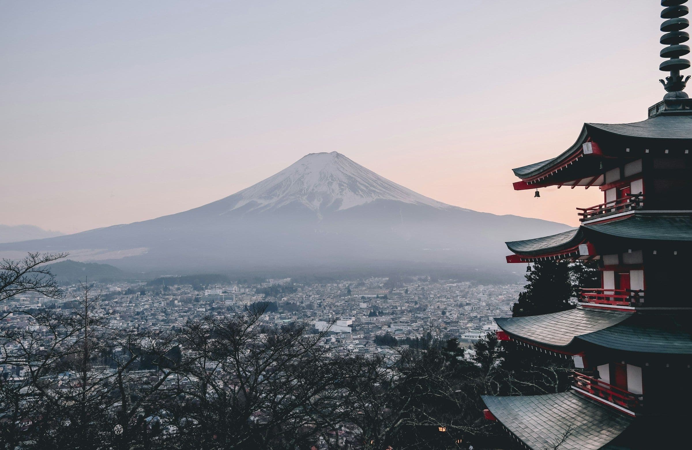 A traditional Japanese pagoda with tiered roofs stands on a mountainside. Mount Fuji, a snow-capped volcano, rises in the background.