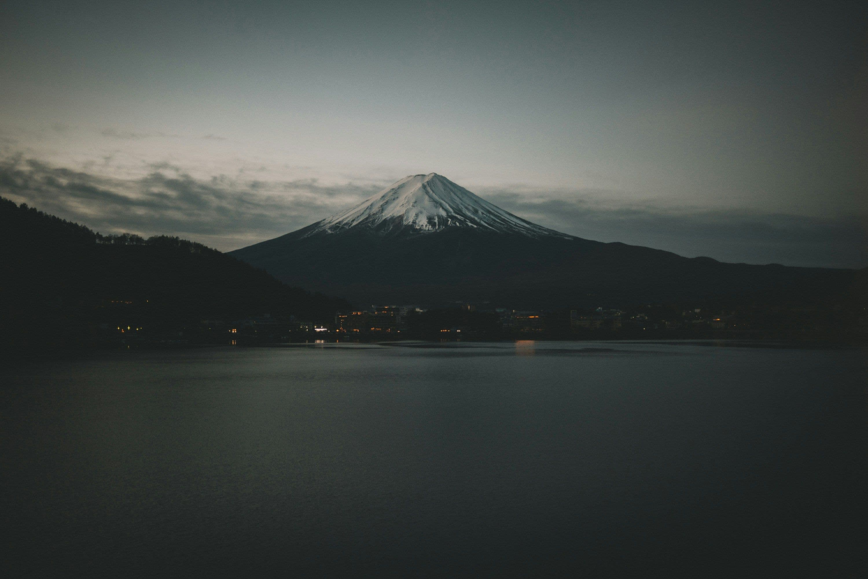 A snow-capped mountain, Mount Fuji, rises above a still lake at dusk. The sky is ablaze with orange, pink, and purple hues.