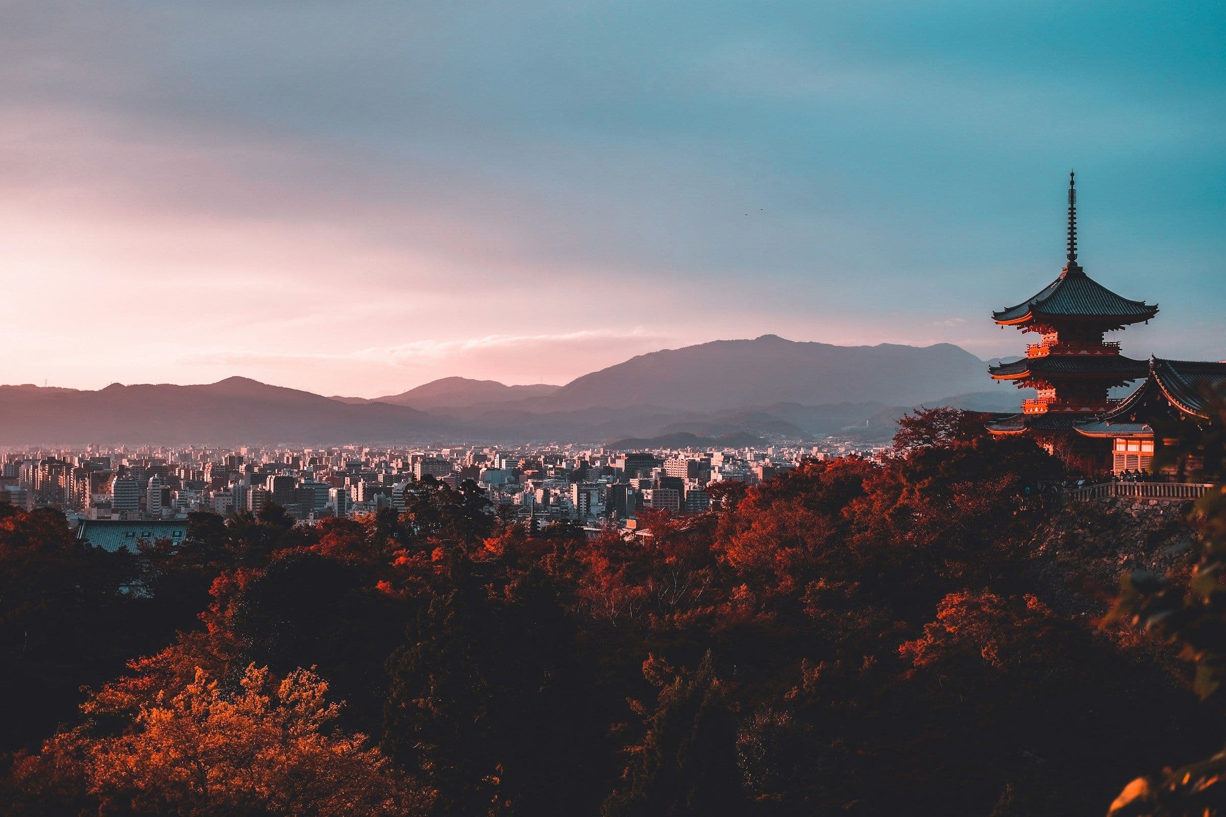 A traditional Japanese pagoda with tiered roofs silhouetted against a colorful sunset. The pagoda overlooks a sprawling city with skyscrapers and high-rise buildings illuminated in the twilight.